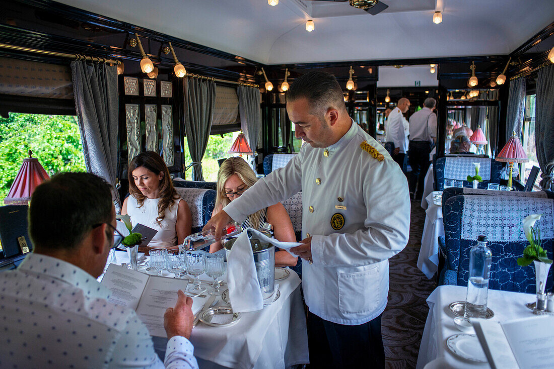 Ein Kellner serviert Wasser und Essen im Art-Deco-Restaurantwagen des Luxuszuges Belmond Venice Simplon Orient Express