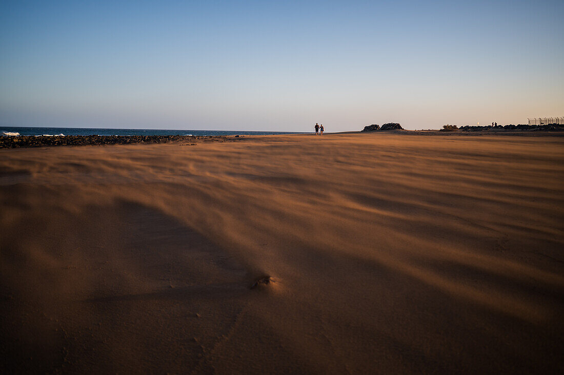 Ein Paar geht am Strand spazieren, während ein starker Wind Sand auf die Insel Lanzarote, Kanarische Inseln, Spanien, weht