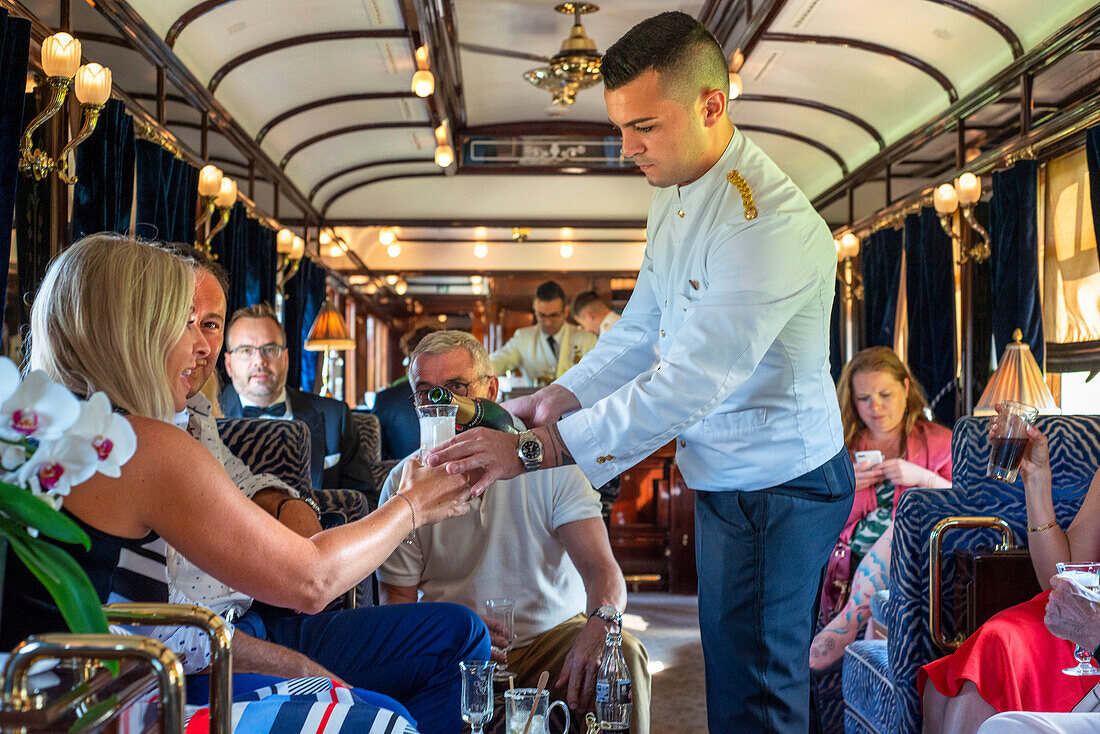 A waiter serves champagne inside the art deco bar lounge wagon of the train Belmond Venice Simplon Orient Express luxury train.