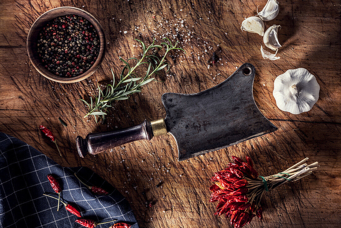 Old butcher's cleaver on a chopping board, surrounded by chilli peppers, garlic and rosemary
