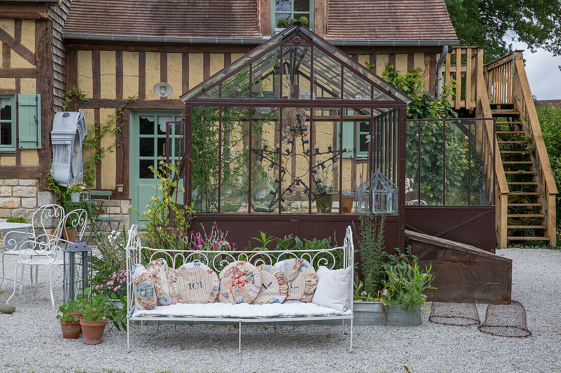 Glasgewächshaus und schmiedeeiserne Sitzbank mit Kissen im Landhausgarten