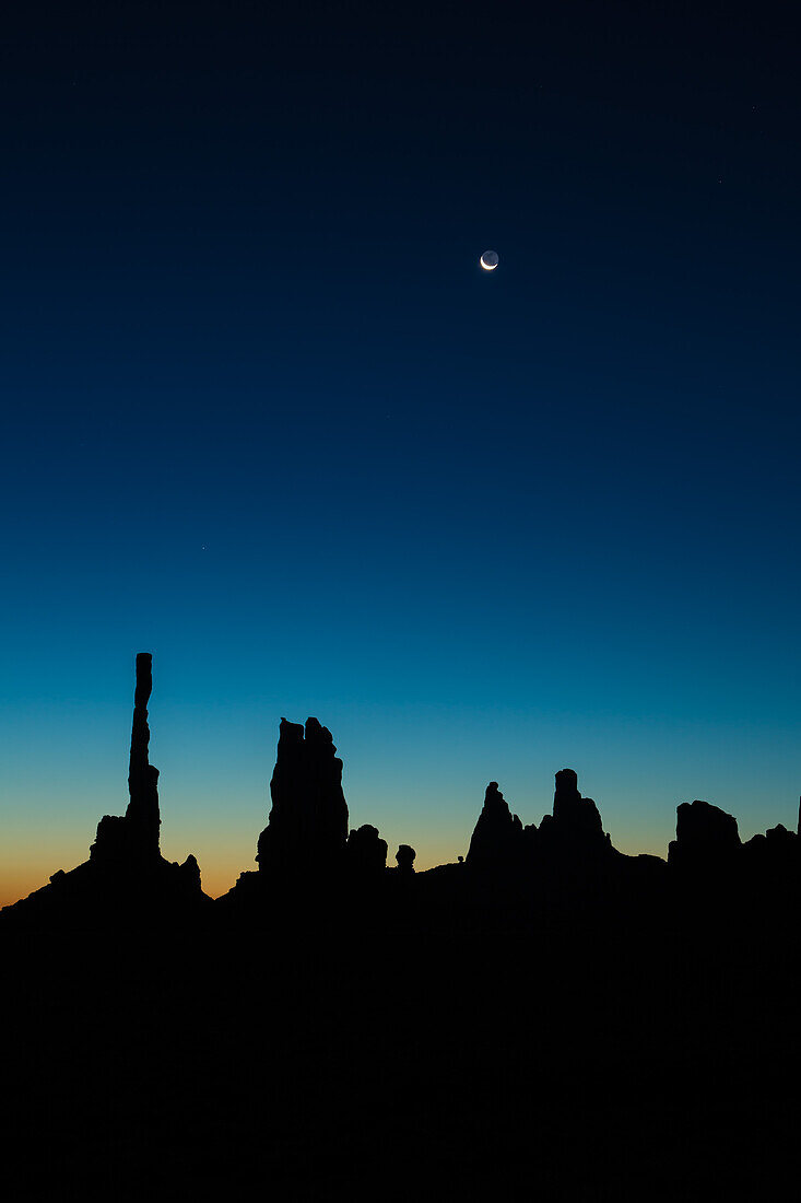 Moon over the Totem Pole and the Yei Bi Chei in silhouette before dawn in the Monument Valley Navajo Tribal Park in Arizona.