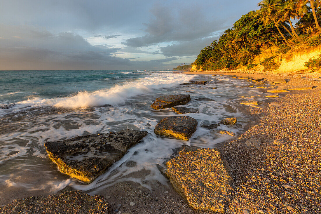 Wellen, die sich bei Sonnenaufgang an einem Strand bei Barahona in der Dominikanischen Republik an den Felsen brechen. Eine lange Verschlusszeit verleiht dem Wasser einen verschwommenen Look