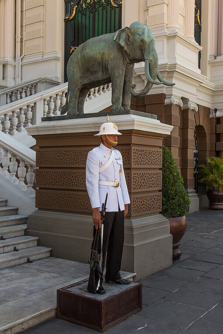 A Thai soldier in ceremonial uniform on duty at the Grand Palace complex in Bangkok, Thailand. An elephant statue is behind.