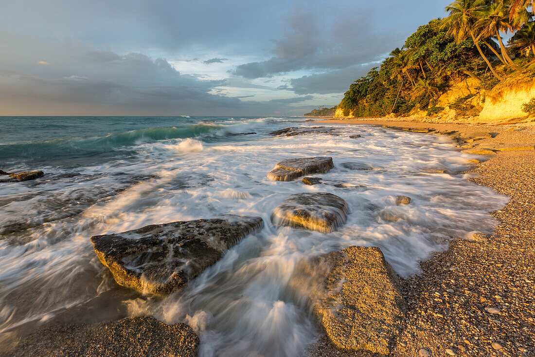 Wellen, die sich bei Sonnenaufgang an einem Strand bei Barahona in der Dominikanischen Republik an den Felsen brechen. Eine lange Verschlusszeit verleiht dem Wasser einen verschwommenen Look