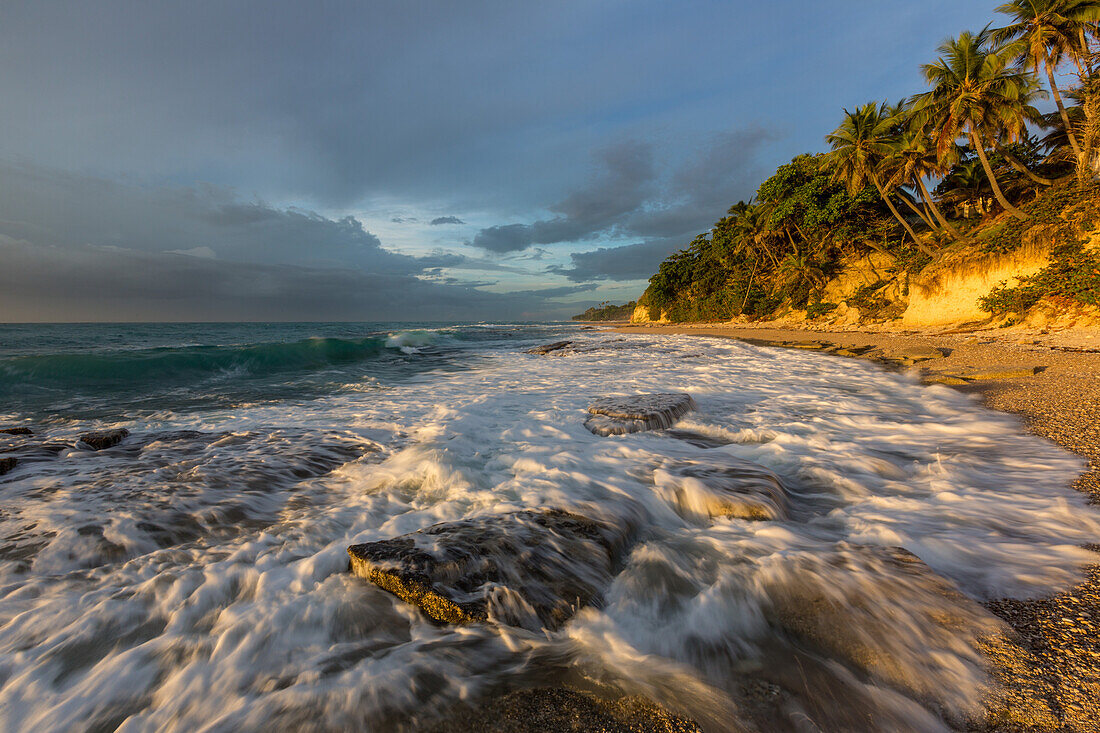 Wellen, die sich bei Sonnenaufgang an einem Strand bei Barahona in der Dominikanischen Republik an den Felsen brechen. Eine lange Verschlusszeit verleiht dem Wasser einen verschwommenen Look