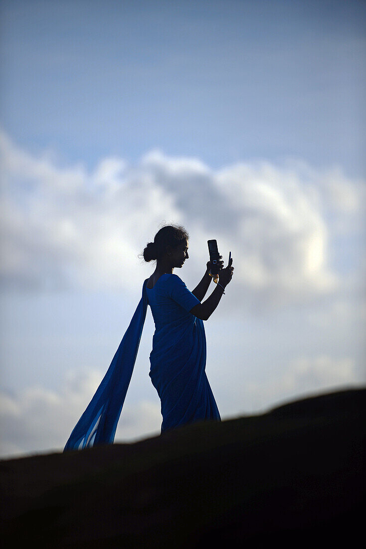 Young woman in numerous group of college students, dressed in blue, uses mobile phone during visit to UNESCO World Heritage, Galle Fort, during Binara Full Moon Poya Day.