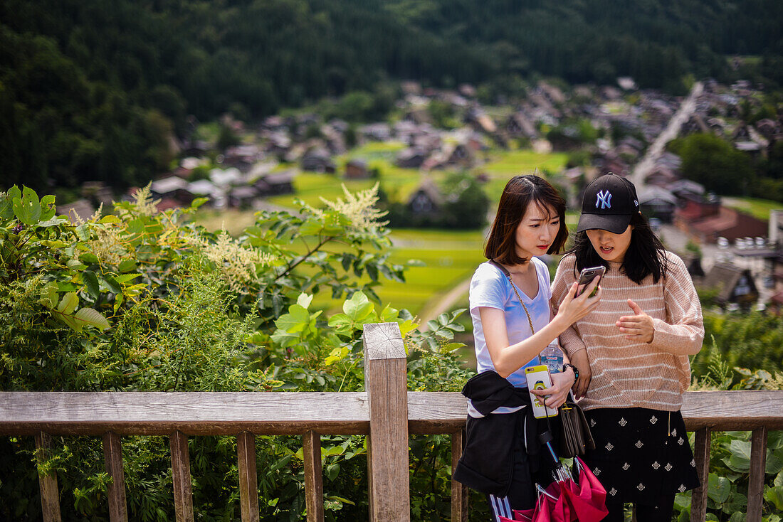 Zwei Mädchen machen ein Selfie von einem Aussichtspunkt in Shirakawa-go, einem traditionellen Dorf, das einen als Gassho-Zukuri bekannten Baustil zeigt, Präfektur Gifu, Japan
