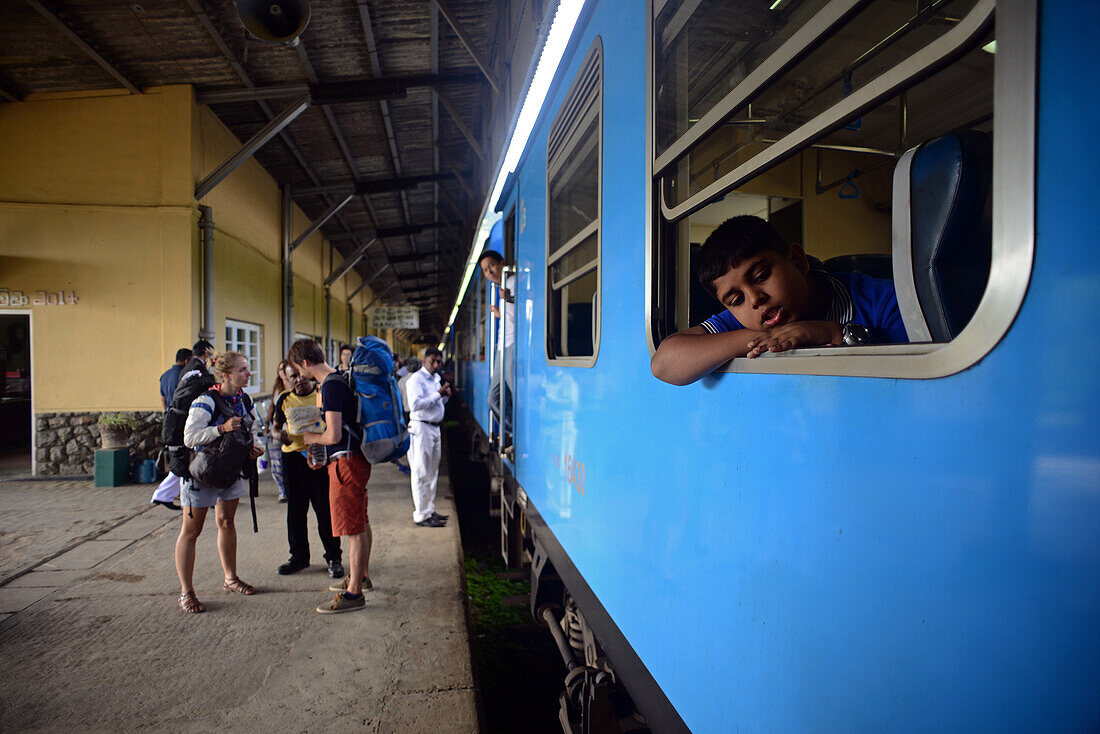 Junger Junge am Fenster, Rucksacktouristen auf dem Bahnsteig und andere Menschen auf dem Bahnsteig. Zugfahrt von Kandy nach Nuwara Eliya, Sri Lanka