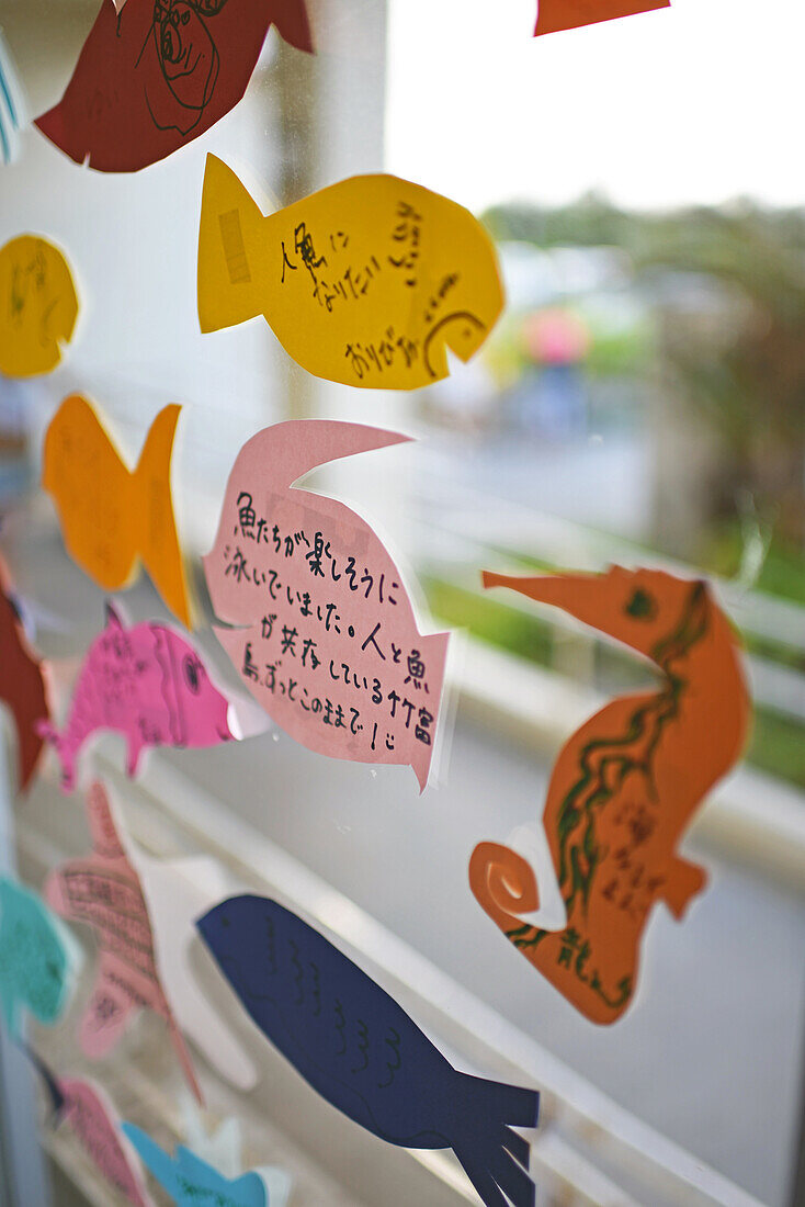 Von Kindern gebastelte Zettel in Fischform schmücken die Glasfenster im Hafen von Taketomi, Okinawa, Japan