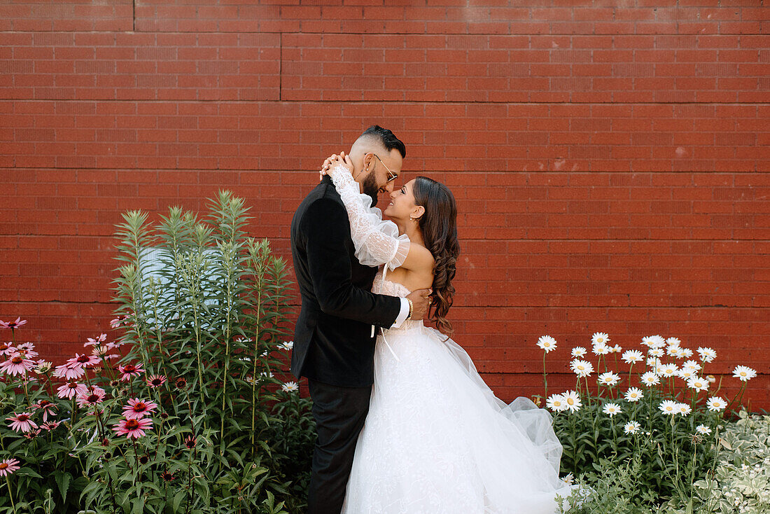 Bride and groom embracing against brick wall and flowers