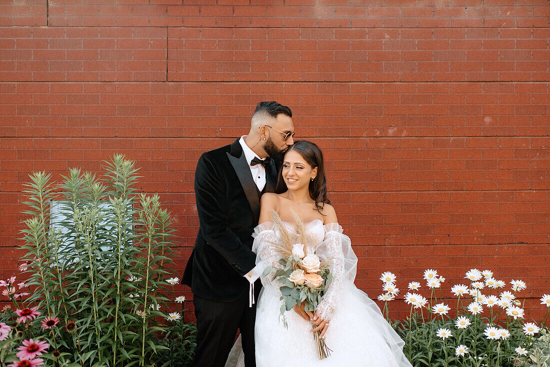 Bräutigam küsst Braut vor einer Mauer und Blumen