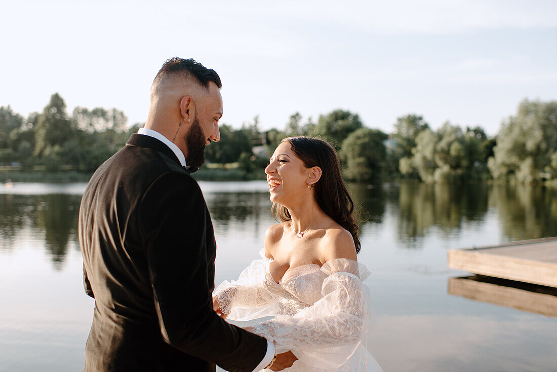 Braut und Bräutigam halten sich am Seeufer an den Händen und lachen