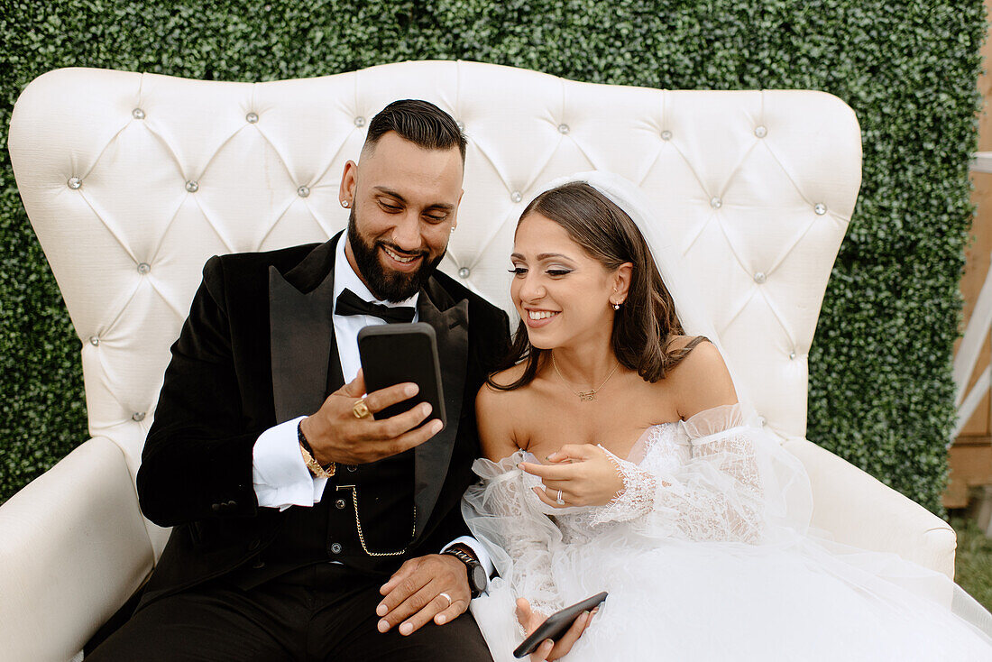 Smiling bride and groom sitting on sofa, looking at smart phone
