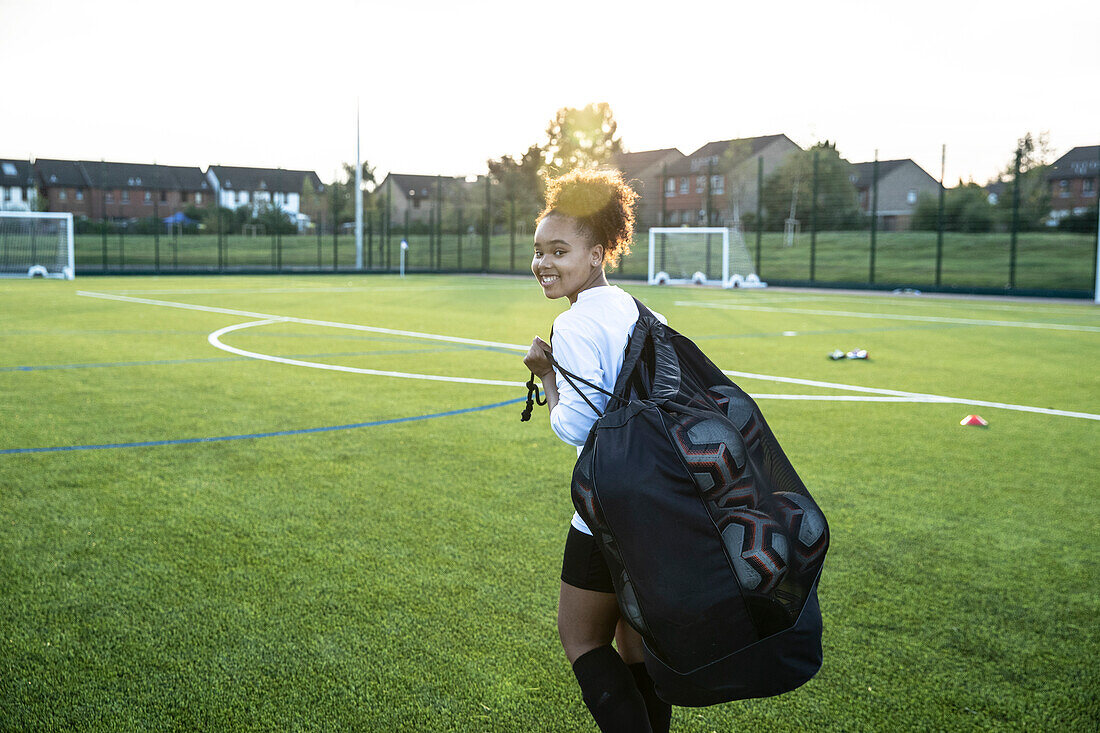 UK, Smiling female soccer player (12-13) carrying bag with balls in field