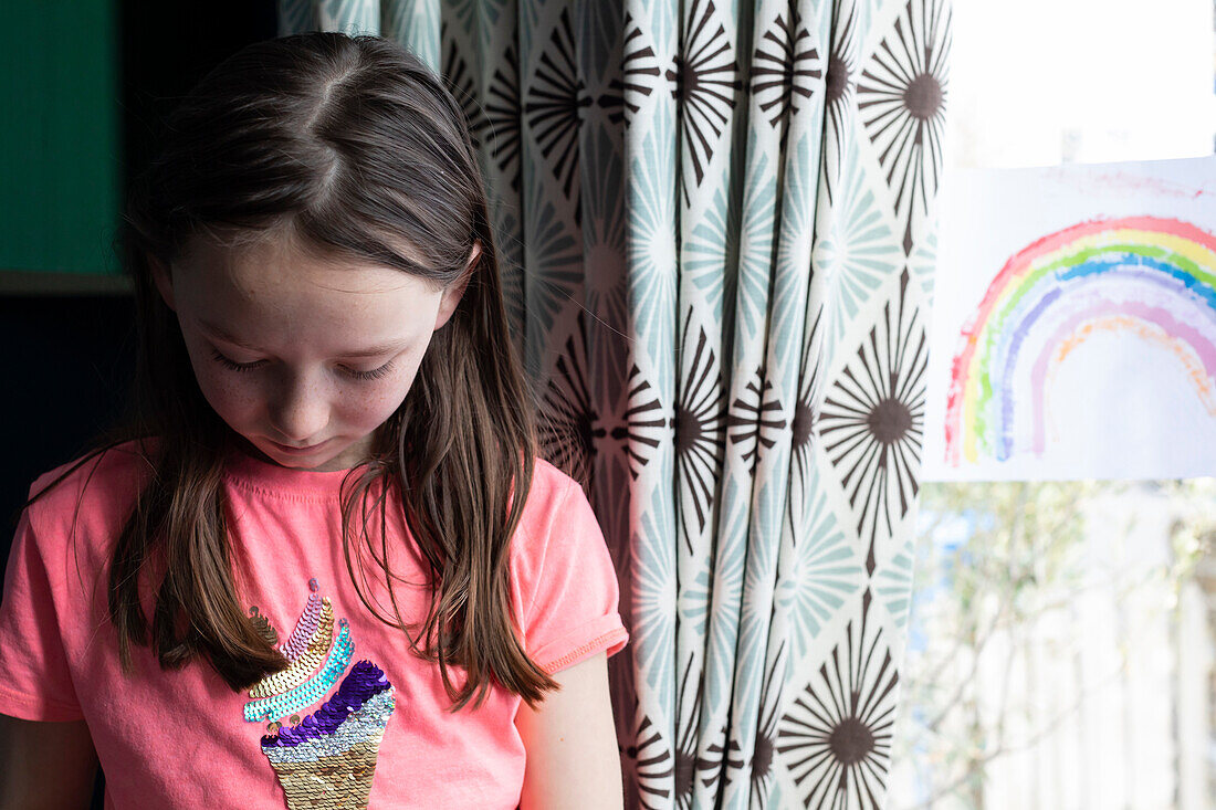 Girl (4-5) by window with drawing of rainbow