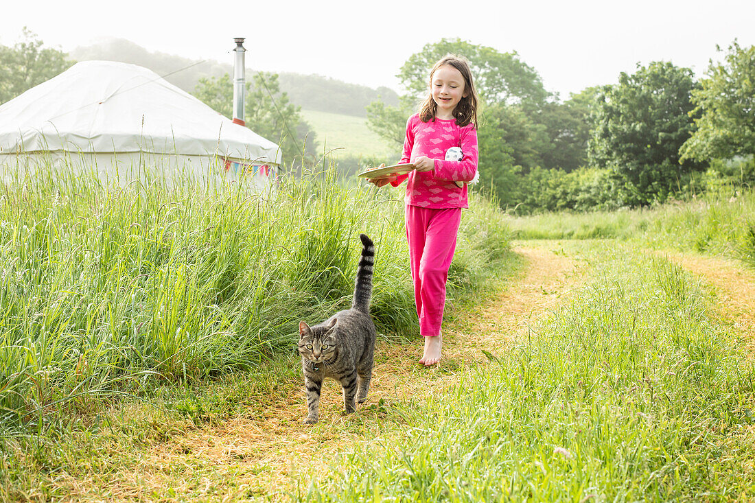 Girl (4-5) in pajamas walking in grassy field with cat near yurts in campsite