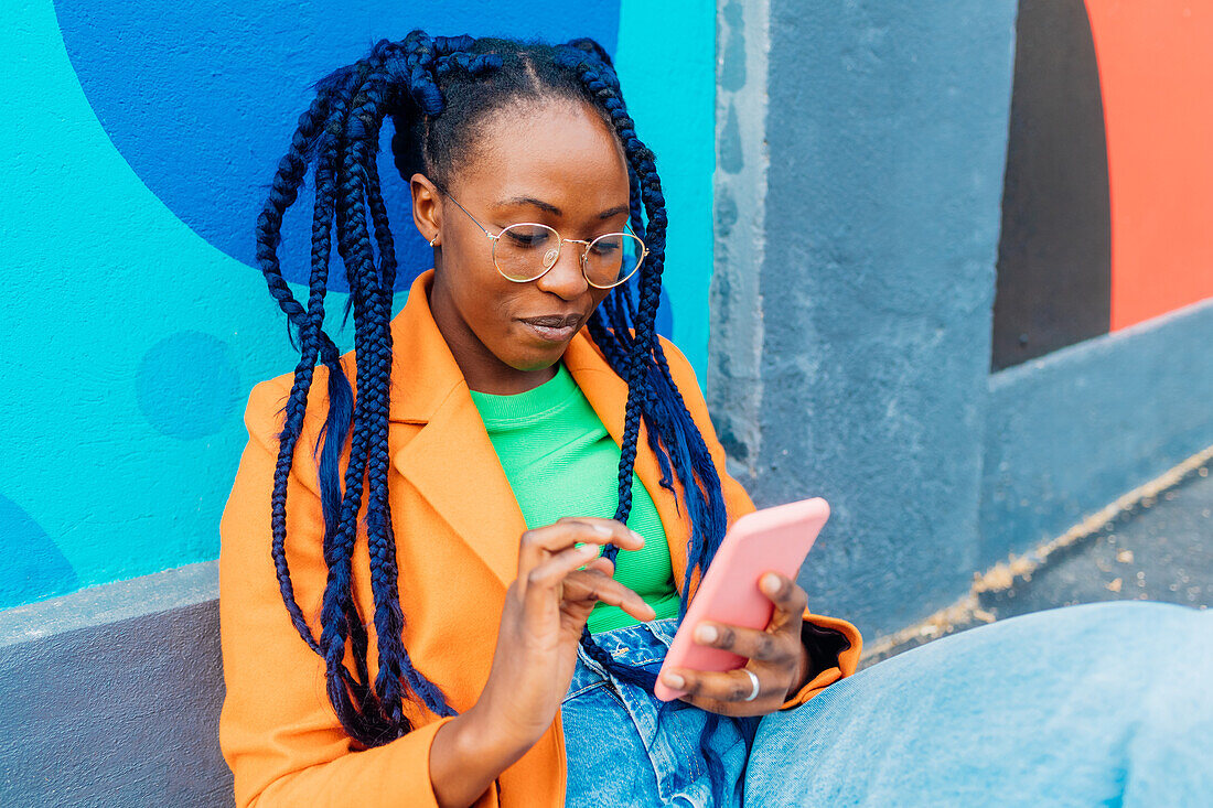 Italy, Milan, Woman with braids sitting by colorful wall, using smart phone