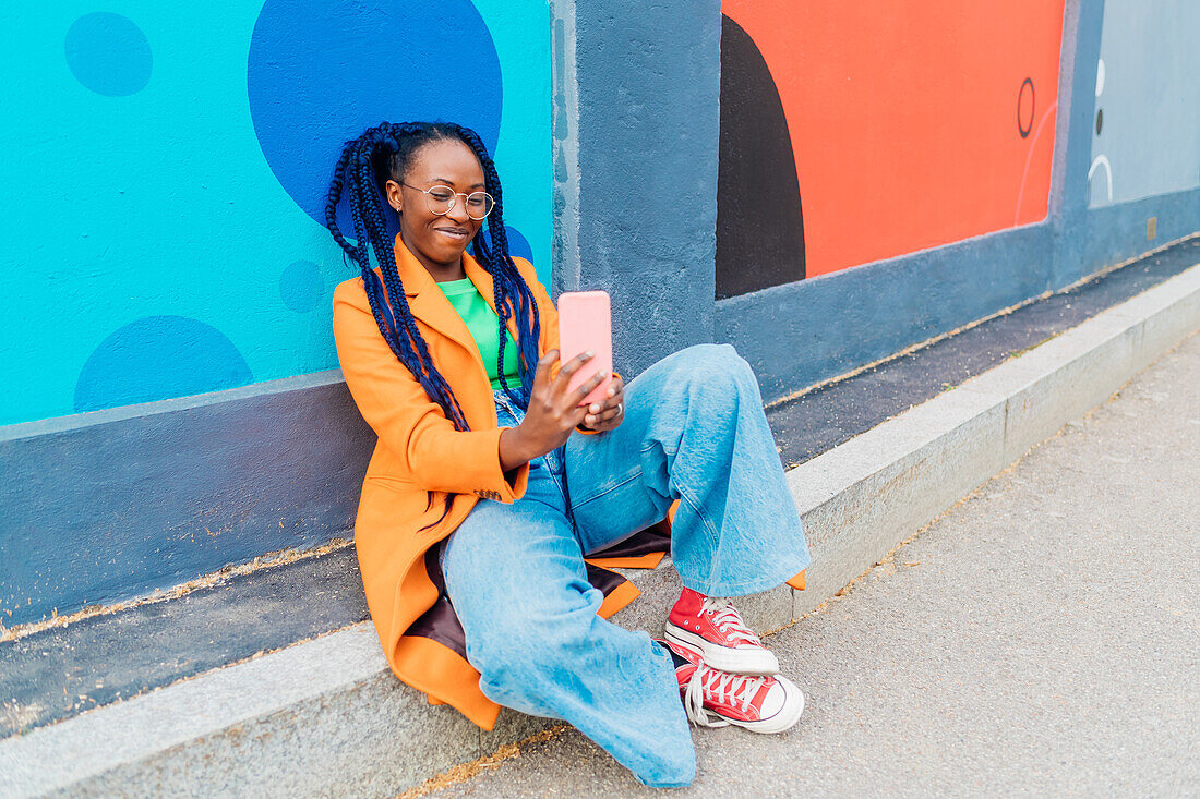 Italy, Milan, Woman with braids sitting by colorful wall, using smart phone