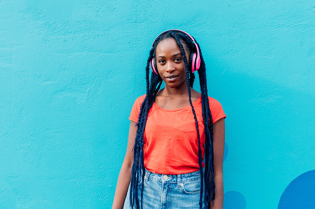 Italy, Milan, Portrait of young woman with headphones in front of blue wall