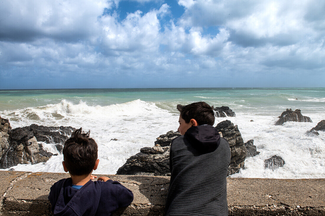 Boys (6-7, 12-13) looking at sea waves crashing against rocks, Sicily, Italy