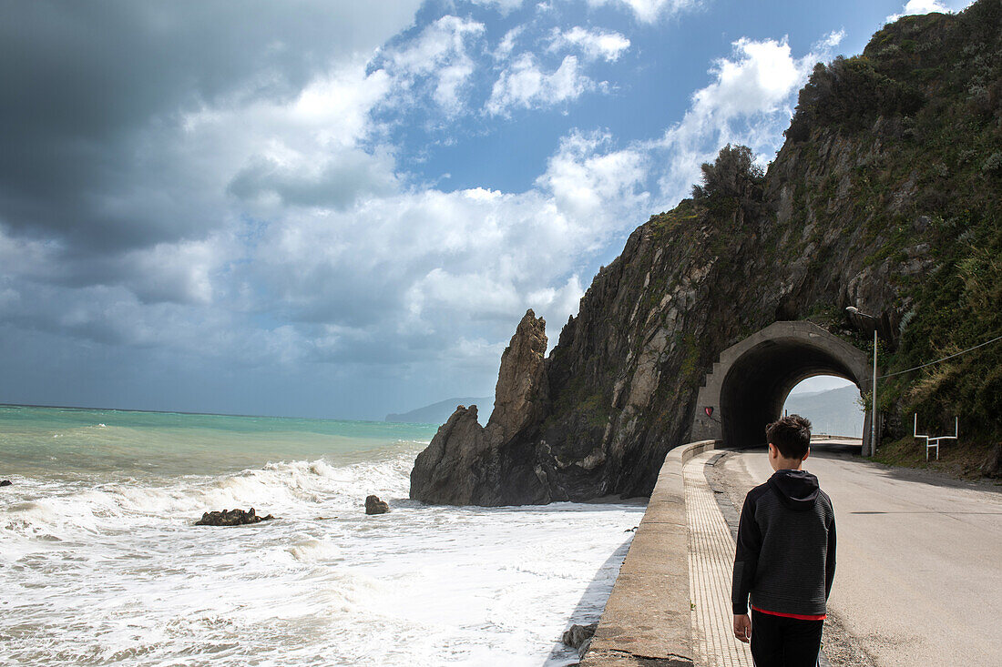 Rear view of boy (12-13) standing on coastal road, Sicily, Italy