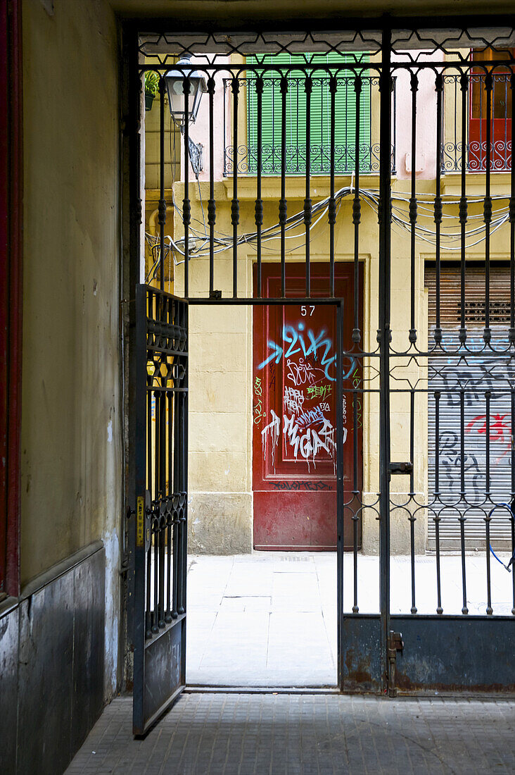 A Metal Gate Open In The Lower Floor Of A Residential Building With Graffiti Painted On The Door Across The Street; Barcelona, Spain