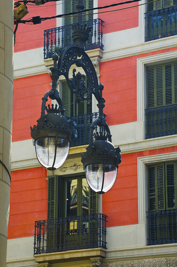 An Ornate Metal Light Fixture Hanging In Front Of A Red Residential Building; Barcelona, Spain