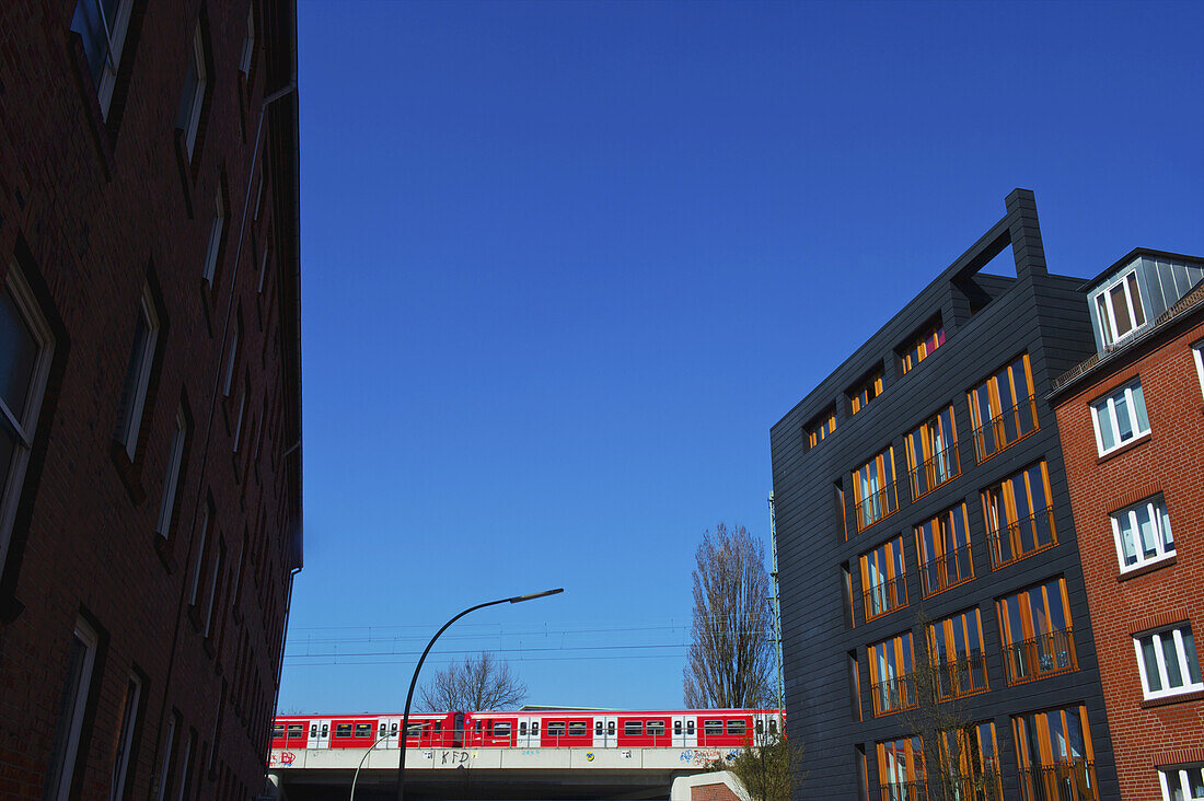 Ein Zug, der über ein erhöhtes Gleis fährt, und ein Wohnhaus aus Backstein mit blauem Himmel; Hamburg, Deutschland
