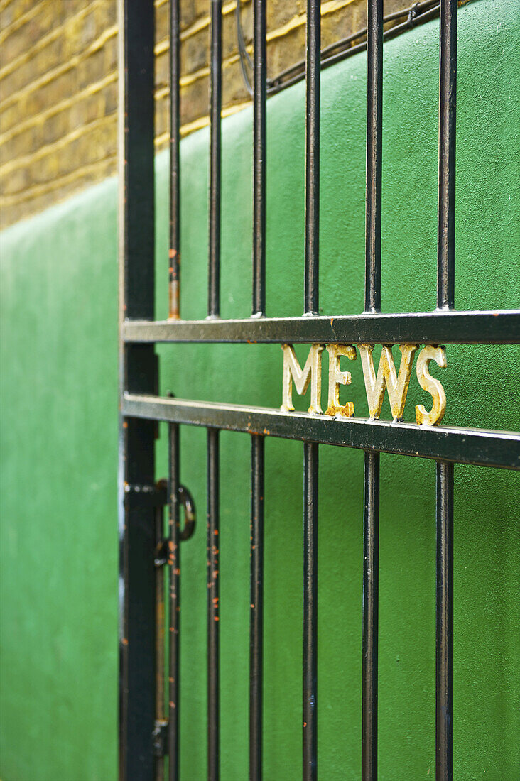 Ein offenes Metalltor mit dem Wort Mews" in goldener Schrift vor einer grünen Wand; London, England".