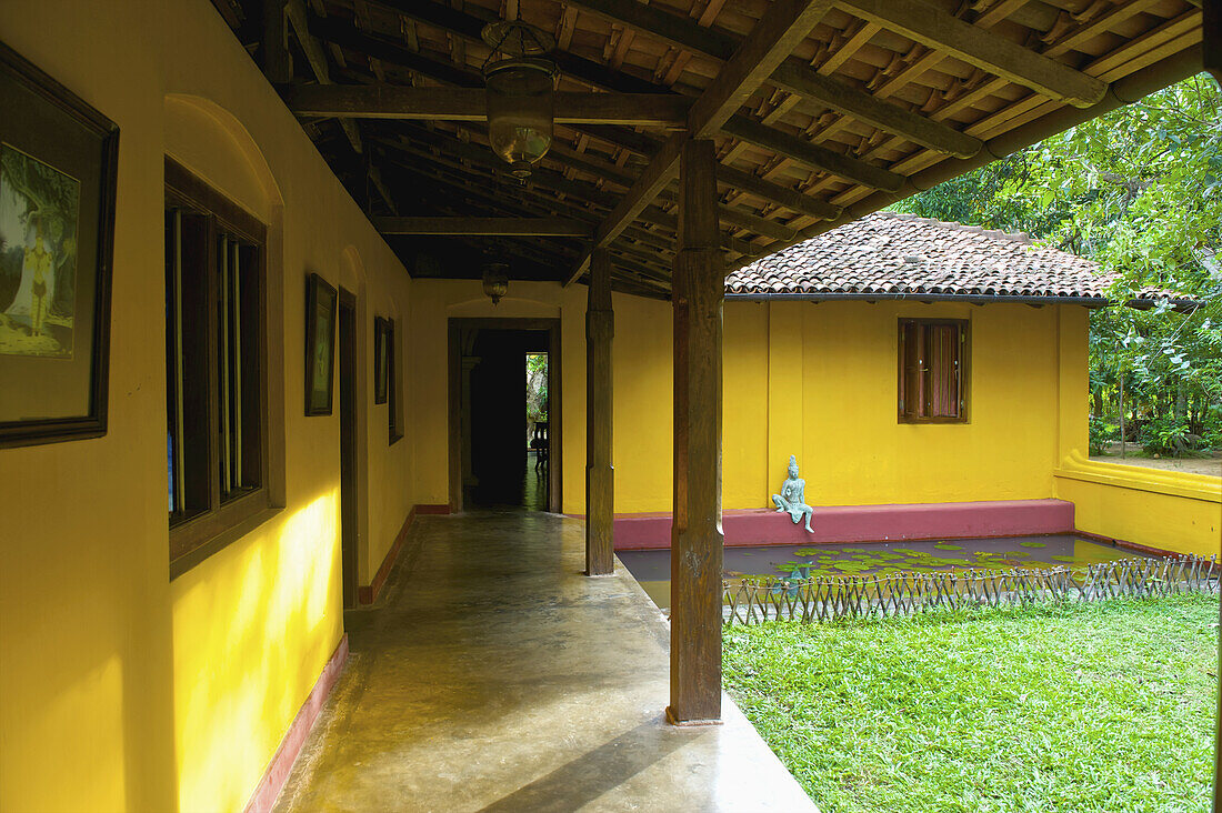 A Bright Yellow Building With A Statue Sitting On A Red Ledge By A Pond; Ulpotha, Embogama, Sri Lanka
