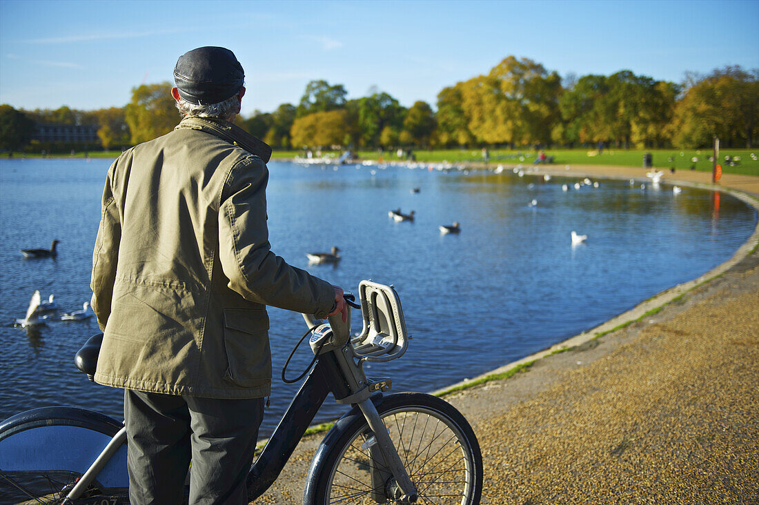 Ein Mann steht mit seinem Fahrrad am Rande des Serpentine Lake, Kensington Gardens; London, England.