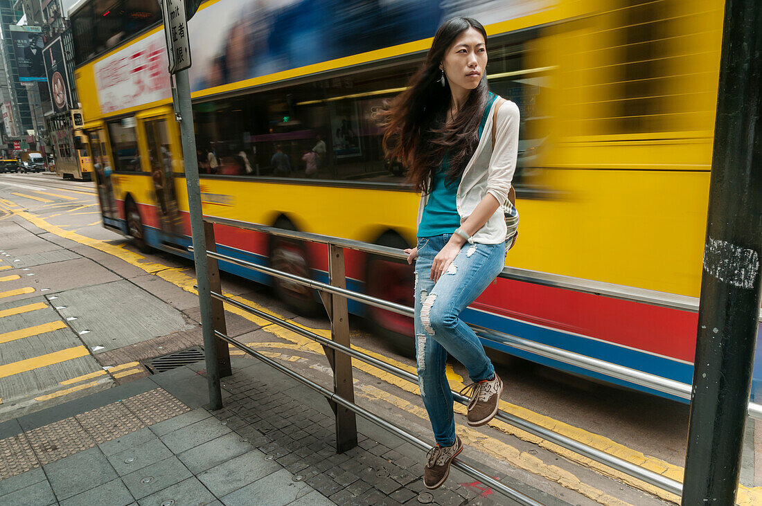 Chinesische junge Frau sitzt auf einem Geländer in einer Straße, hinter ihr fährt ein zweistöckiger Bus; Hongkong, China