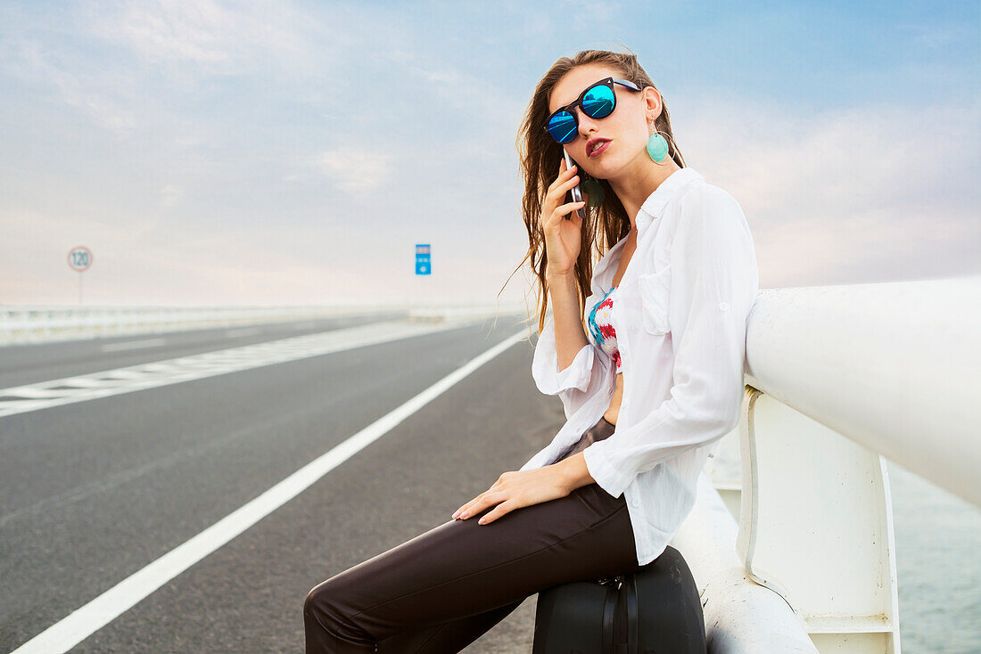 Frau telefoniert mit ihrem Mobiltelefon am Rande einer Autobahn, müde sitzt sie in ihrem Gepäck; Xiamen, Fujian, China