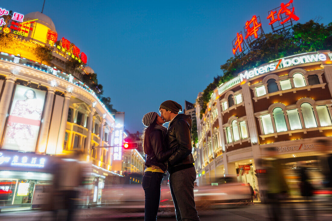 A Couple Kissing With The Blur Of Traffic And Pedestrians In The Background; Hong Kong, China