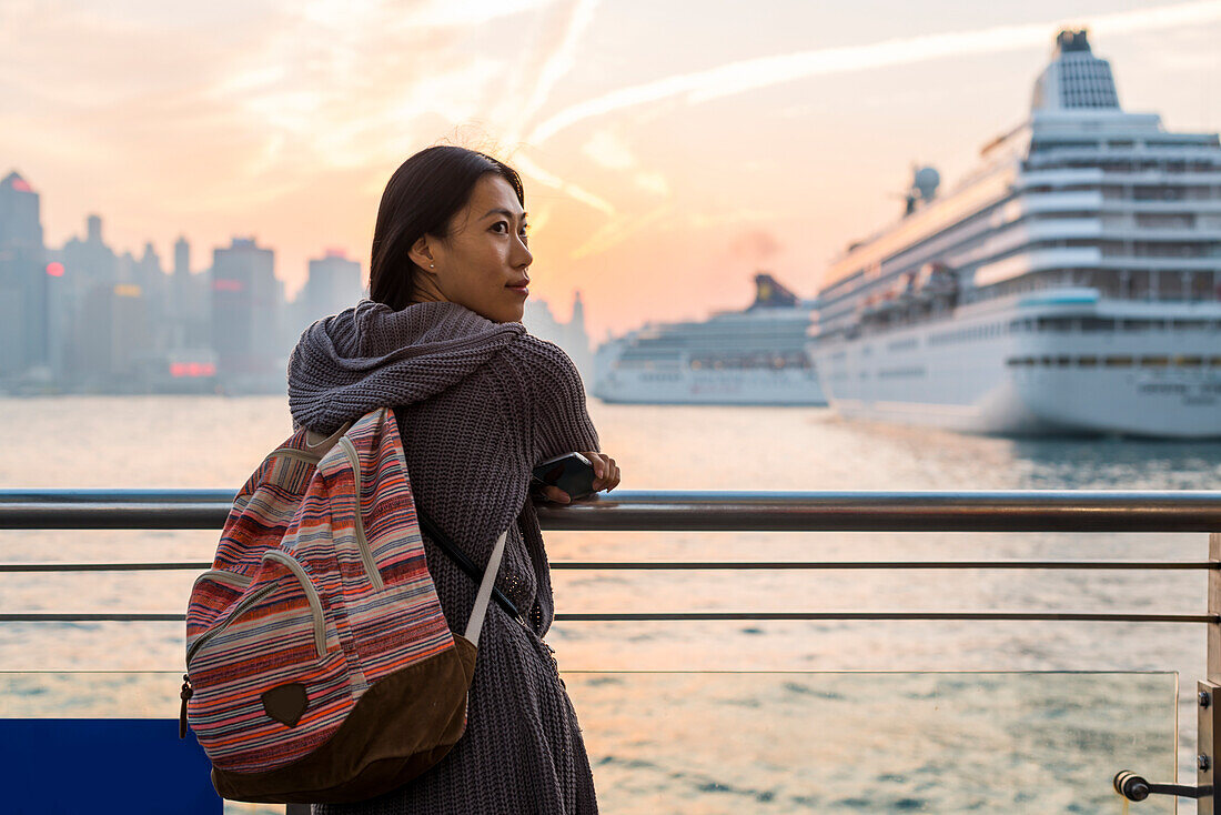 Junge Frau an der Uferpromenade mit Kreuzfahrtschiffen im Hafen im Hintergrund, Kowloon; Hongkong, China.