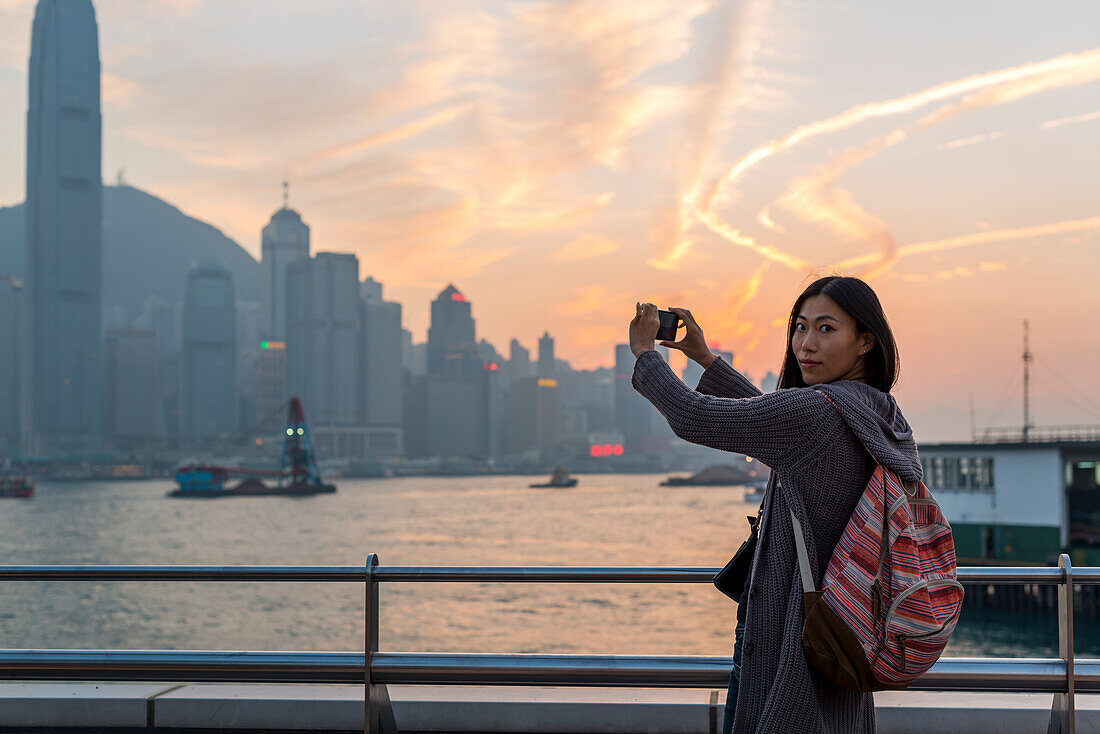 Eine junge Frau am Wasser mit Blick auf die Skyline bei Sonnenuntergang, Kowloon; Hongkong, China.