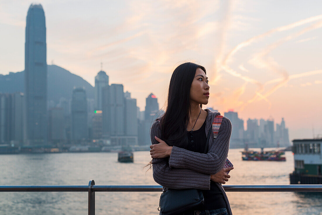 Eine junge Frau am Hafen mit Blick auf die Skyline bei Sonnenuntergang, Kowloon; Hongkong, China.