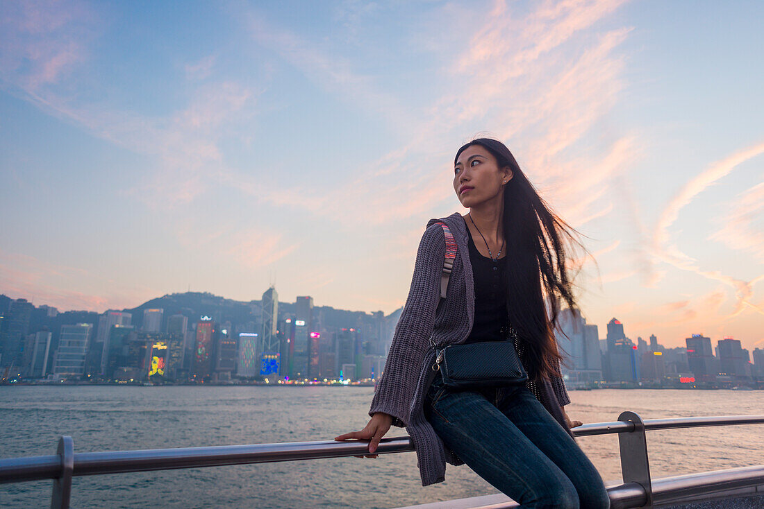Eine junge Frau am Ufer, die bei Sonnenuntergang auf dem Geländer sitzt, mit der Skyline im Hintergrund, Kowloon; Hongkong, China.
