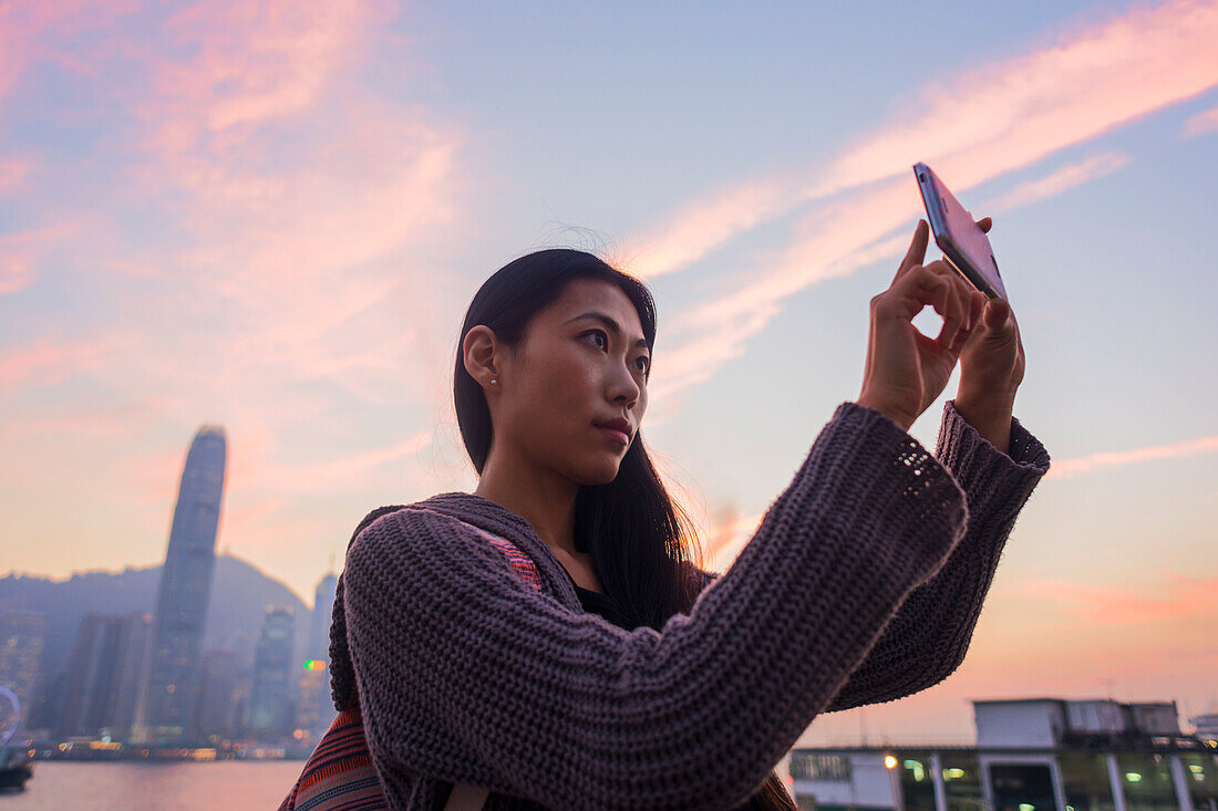 Eine junge Frau am Ufer, die bei Sonnenuntergang ein Foto mit der Skyline im Hintergrund macht, Kowloon; Hongkong, China.