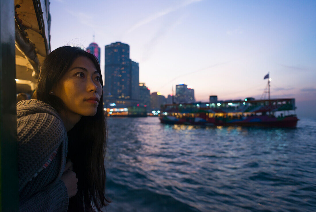 Eine junge Frau am Hafen bei Sonnenuntergang mit einem Boot und der Skyline im Hintergrund, Kowloon; Hongkong, China.