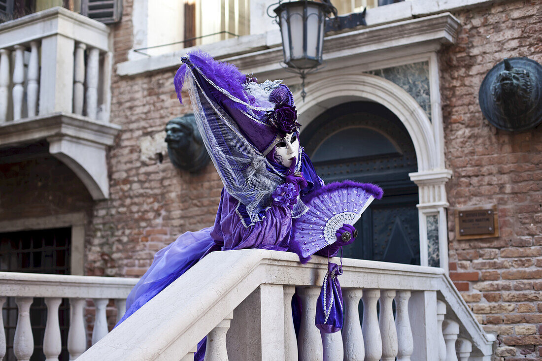 Eine Frau in einem lila Kleid und Fächer, posierend an einem Gebäude und einem Geländer; Venedig, Italien.