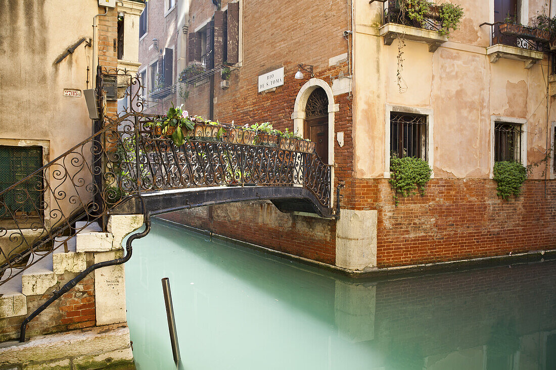 Footbridge With Ornate Railing And Decorative Plants Crossing A Canal; Venice, Italy