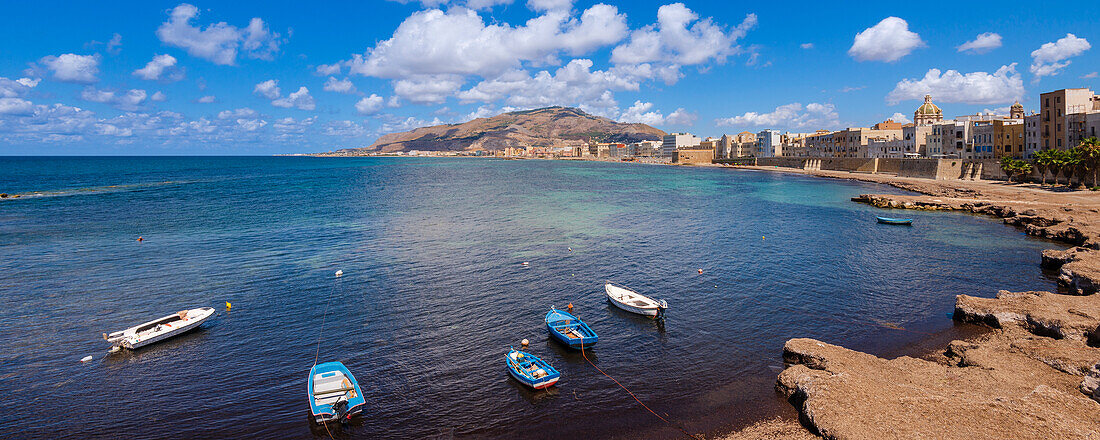 Small boats moored along the shore of the Trapani skyline with old, stone buildings and sea wall; Trapani, Sicily, Italy