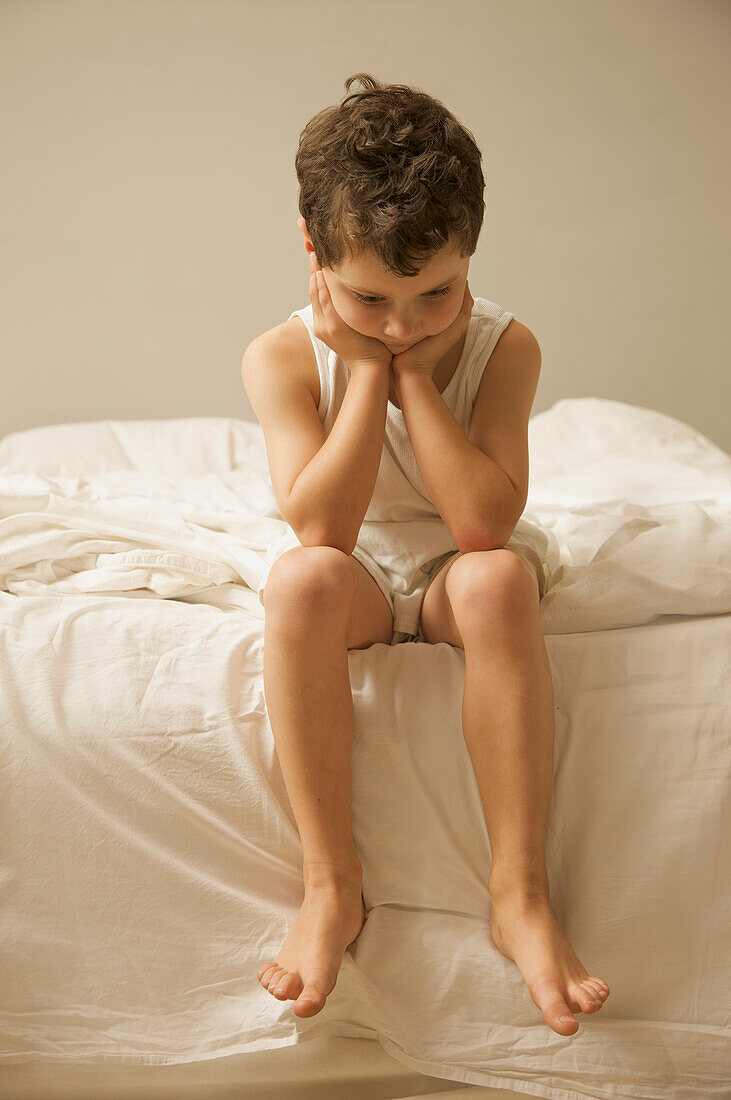 Young boy sitting on the end of a bed with face resting on hands