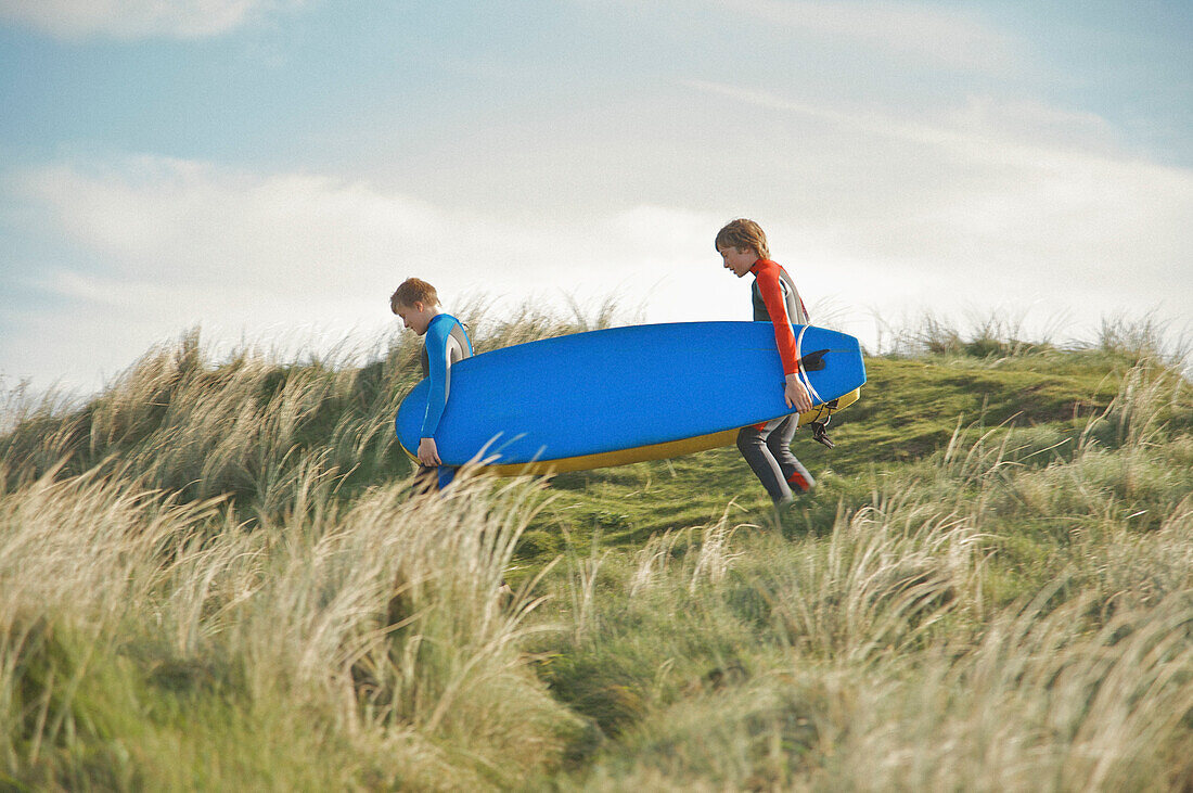 Two teenaged boys walking on a sand dune holding surfboards