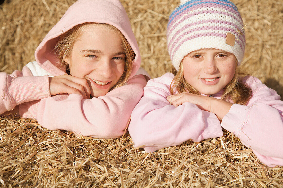 Two young girls leaning on bale of hay smiling