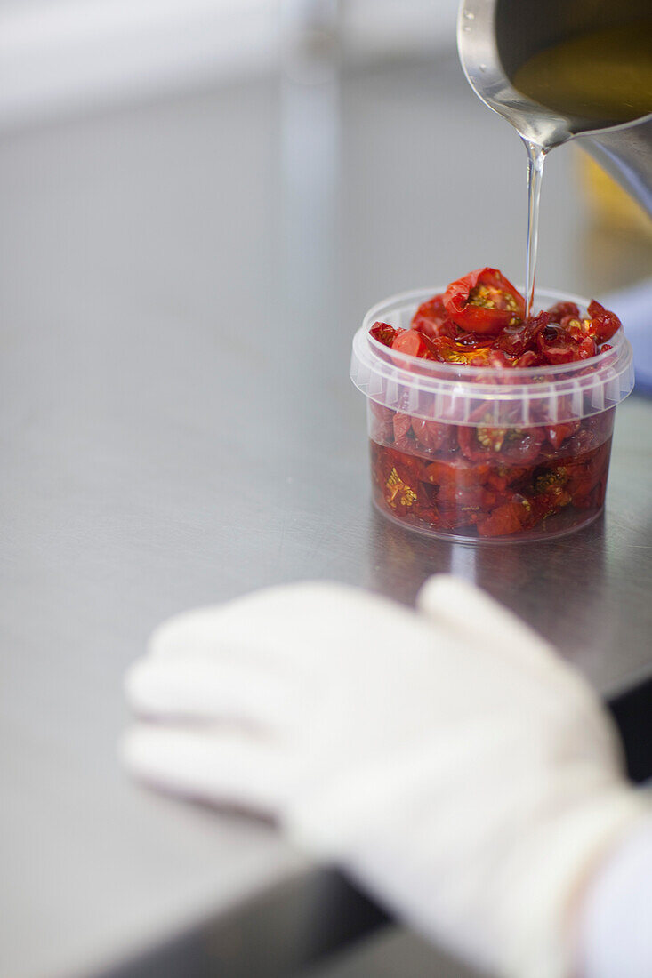 Nahaufnahme eines Arbeiters, der mit der Hand Öl in einen mit sonnengetrockneten Tomaten gefüllten Kunststoffbehälter gießt