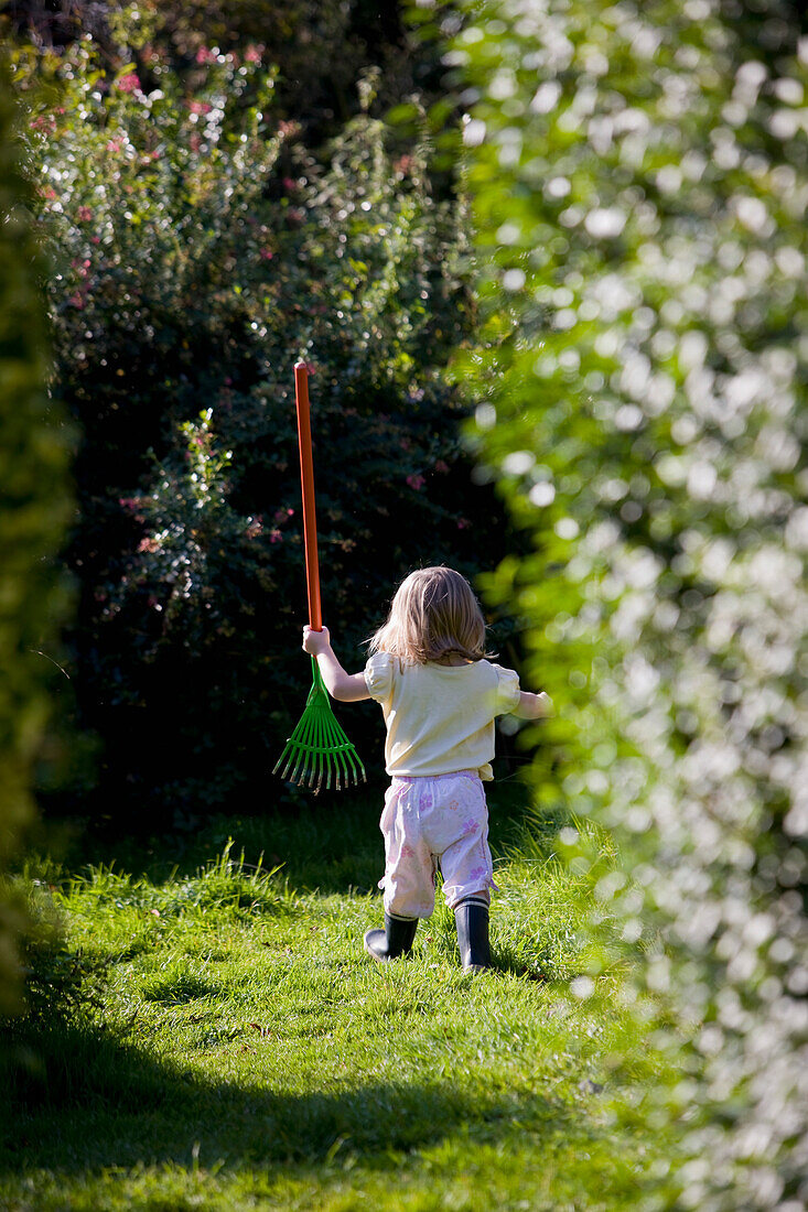 Back view of a young girl walking in a garden holding a toy rake