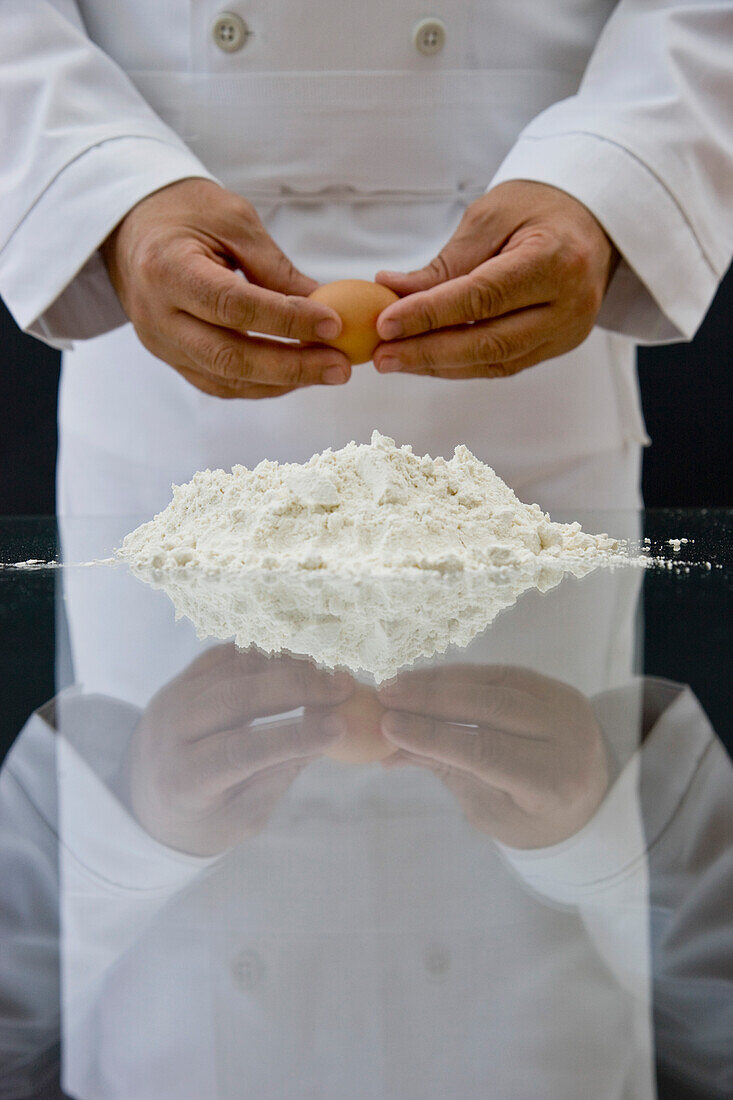 Nahaufnahme eines Kochs, der mit seinen Händen ein Ei über einem Mehlhaufen auf einem Glastisch hält